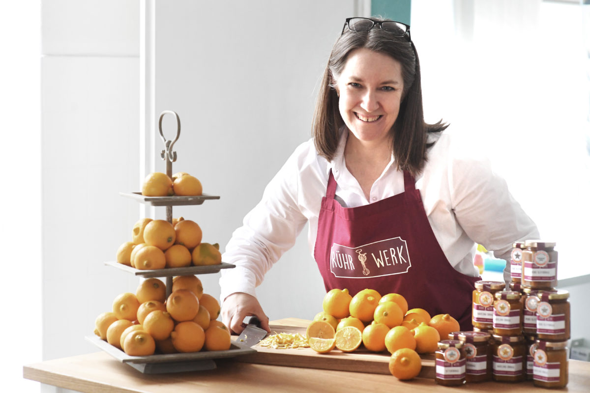 Frau Rührwerk mit Schürze und Messer um Orangenschalen für die englische Marmalade zu schneiden, mit Agrumen und fertigen Zitrusmarmeladen auf dem Tisch.